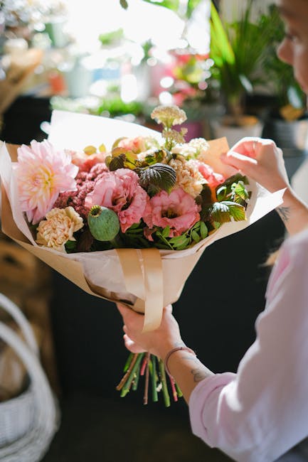 A person holding a bouquet of fresh flowers wrapped in beige paper and tied with a cream ribbon, featuring pink peonies, soft pink carnations, white blossoms, and green foliage. The bouquet is arrange