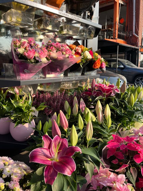 A vibrant display of fresh flowers arranged on outdoor shelving at a flower shop, featuring pink and white lilies with closed buds, bright pink poinsettias, and small bouquets of mixed pink, white, an