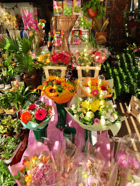 A vibrant display of fresh floral arrangements and potted plants inside a flower shop, featuring bouquets of bright yellow, pink, and red flowers wrapped in colorful paper, placed on a pink tablecloth