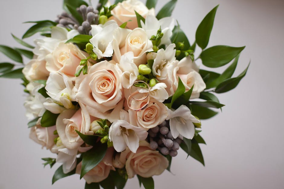 A close-up view of a floral bouquet arranged with pale pink roses, white freesias, and small grey berries, accented by lush green leaves. The roses are fully bloomed with soft, velvety petals, while t