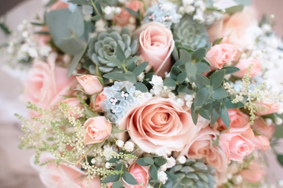 A close-up image of a floral arrangement featuring large, soft pink roses and smaller matching pink rosebuds surrounded by silver-green eucalyptus leaves and delicate white baby's breath. The bouquet