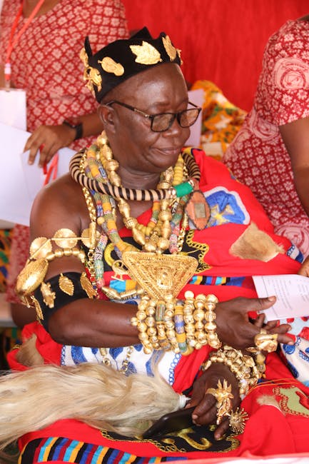 The image features an elderly woman dressed in vibrant traditional African attire adorned with elaborate gold jewelry including necklaces, bracelets, and earrings. She is wearing a black headpiece dec