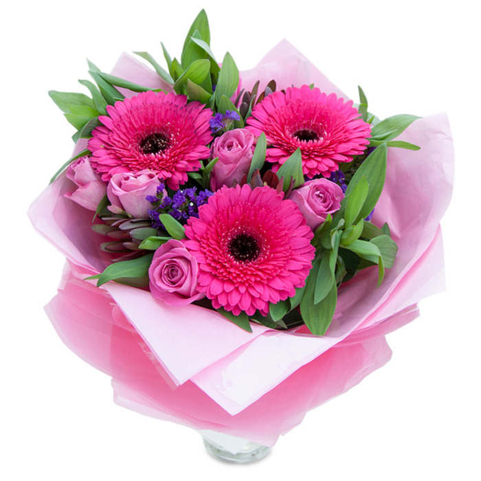 The charm of this pink-focused bouquet lies in the balance of bold and soft elements: three fuchsia Gerbera daisies open wide with confident, layered petals circling deep brown centres that draw the eye, while a collection of lavender-tinged roses offers a softer, dusty-rose counterpoint with petals still gently furled. Between and beneath these focal blooms, clusters of purple statice add a cool, papery texture that punctuates the palette and creates small pockets of visual air. Lush greenery-broad, glossy leaves that read as salal or eucalyptus-forms a rich, supportive backdrop, giving weight and a fresh, slightly dewy scent to the arrangement. The wrapping is considered and deliberate: an inner sheet of matte pale pink tissue cradles the stems, while an outer layer of translucent fuchsia-pink cellophane, faintly crinkled, adds a shimmering, cohesive finish. The visible top rim of a clear glass vase beneath the wrapping signals the bouquet is prepared for immediate display and underscores the freshness of the stems. In gentle daylight the fuchsia becomes jewel-like against the softer rose tones; under lamplight the dusty lavender of the roses takes on a warmer blush. Texturally the bouquet invites touch-the velvety rose petals, the taut Gerbera florets, the crisp leaves and the slightly crackling cellophane-each element carefully chosen and placed. I imagine this piece being delivered across Bellingham to mark birthdays, anniversaries, or to offer thanks, arriving as a neat, confident statement of affection that combines florist skill with a neighbourhood sensibility familiar to those who know the local streets and parks.