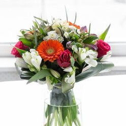 In this artfully arranged bouquet, the essence of Bellingham's friendly neighborhoods comes alive. At its core, velvety orange and creamy peach gerbera daisies glow, their wide petals radiating a gentle energy reminiscent of sunrise over Beckenham Hill. Intertwined among them, the rich magenta roses offer a lush, romantic depth, their tightly curled petals exuding elegance and a hint of drama. Ivory-white alstroemeria and clusters of petite white blossoms are tastefully interspersed, bringing brightness and a light, airy charm to the display. The expertly folded aspidistra leaves, broad and glossy, cradle the arrangement in verdant tranquility, their graceful arcs reflecting the natural form found in Bellingham's garden squares. Smaller, pointed leaves and sprigs of fresh greenery add layers of vitality and structure, framing each bloom and evoking a garden's fullness at the peak of spring. Light softly illuminates the bouquet from a window-bright background, enhancing the vivid hues and delicate textures without overwhelming the senses. Contained in a tall, slender glass vase, the stems are visible-a testament to the bouquet's organic freshness and the florist's eye for detail. The design exudes a sense of optimism, celebration, and new beginnings, making it a perfect offering for local gatherings, housewarmings, or joyful milestones. Every element-down to the last whisper of fragrance-reflects the heartfelt care and expertise of the Bellingham floral community.
